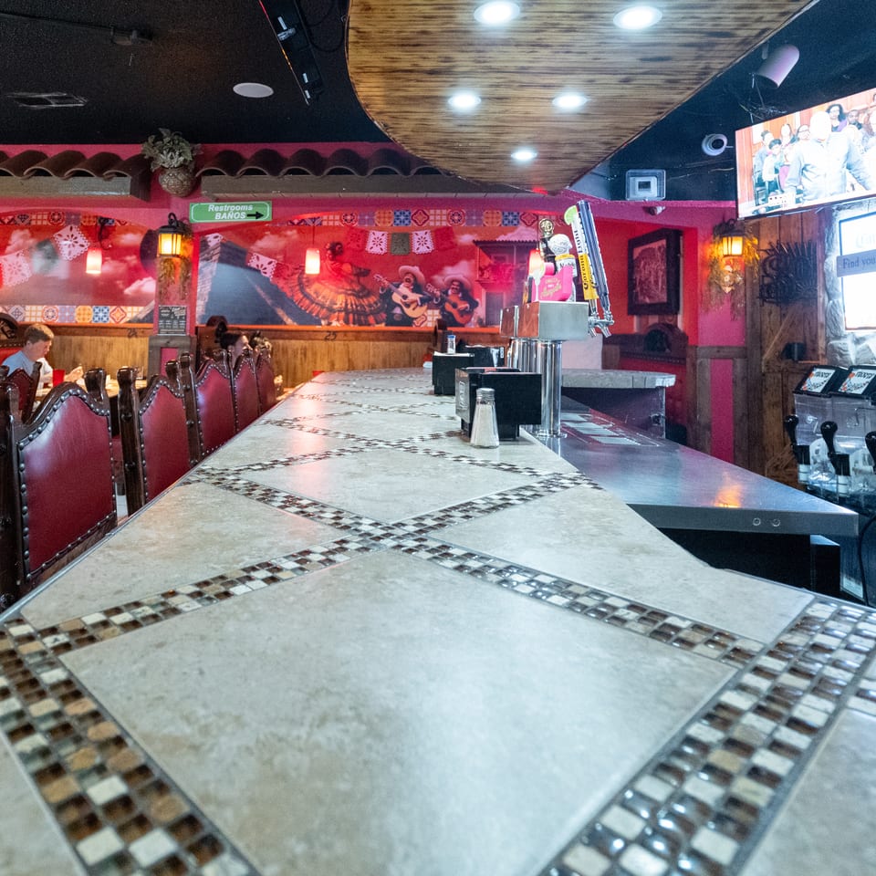 Colorful Mexican restaurant interior with tiled tables, red leather seating, wall murals, hanging lanterns, and a bar area.