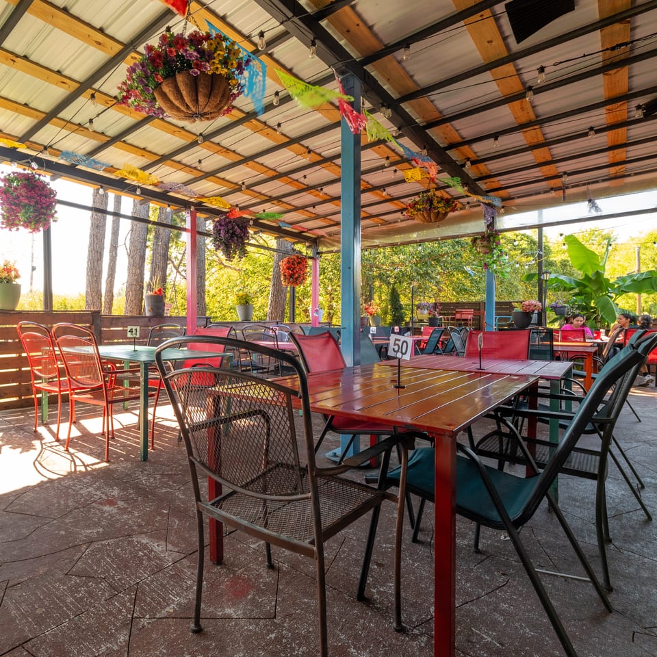 Covered outdoor restaurant patio with colorful hanging flower baskets, red tables and chairs, and diners surrounded by trees.