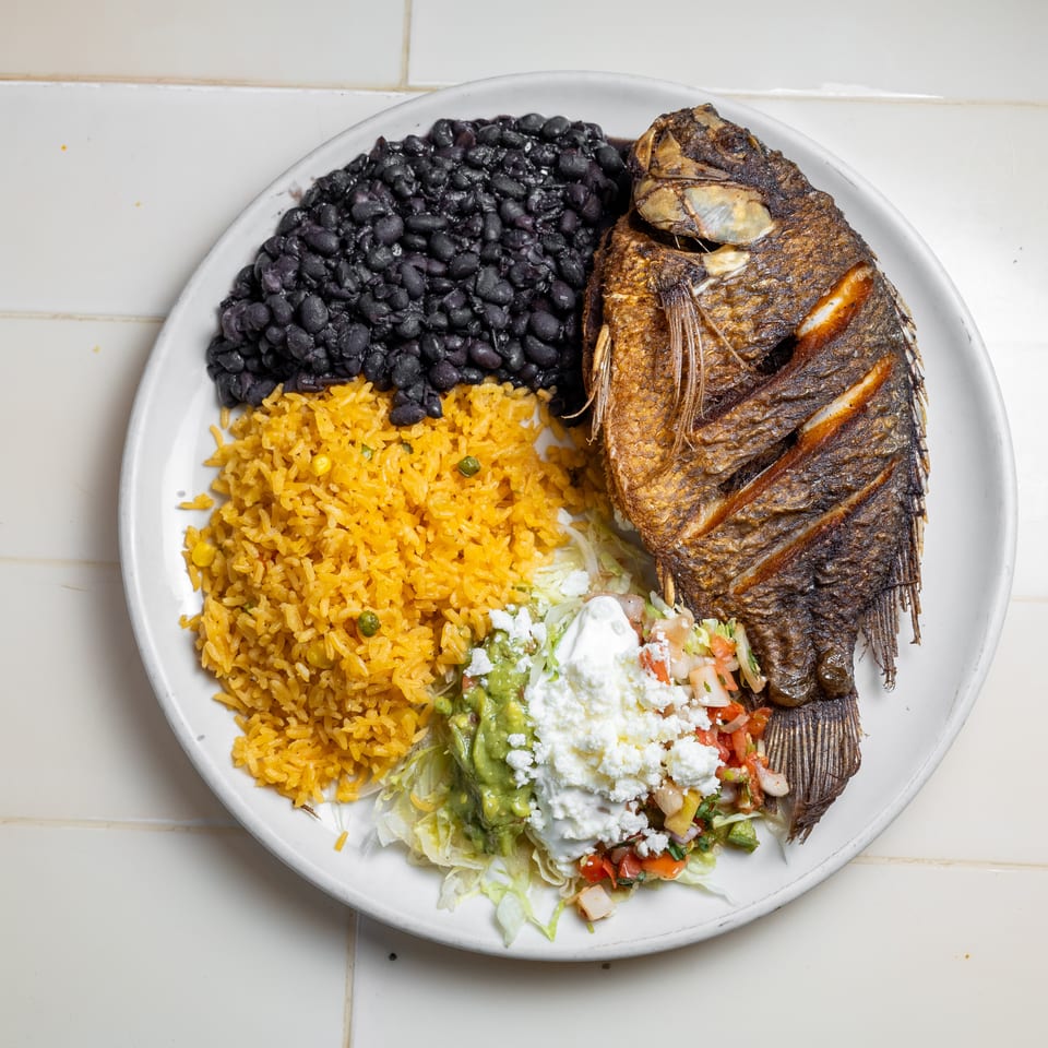 Plate of fried whole fish with black beans, yellow rice, guacamole, salad, pico de gallo, and crema.