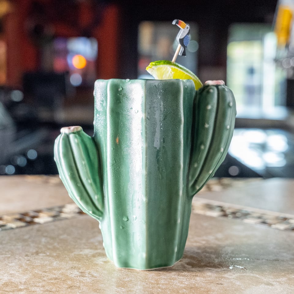 Cactus-shaped green cocktail glass with condensation, lime wedge, and decorative straw on a restaurant bar.