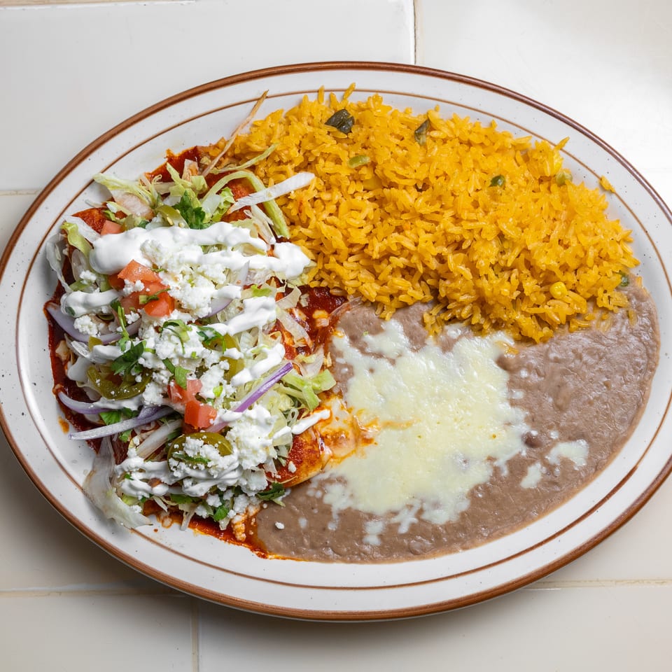 Mexican platter with yellow rice, refried beans topped with melted cheese, and enchiladas covered in lettuce, salsa, crema, and queso fresco.