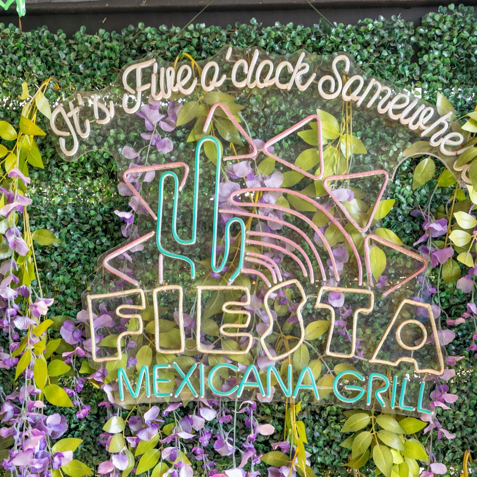 Neon sign reading “Frida & Chaco San Miguel” and “Fiesta Mexicana Grill” surrounded by lush green and purple foliage.