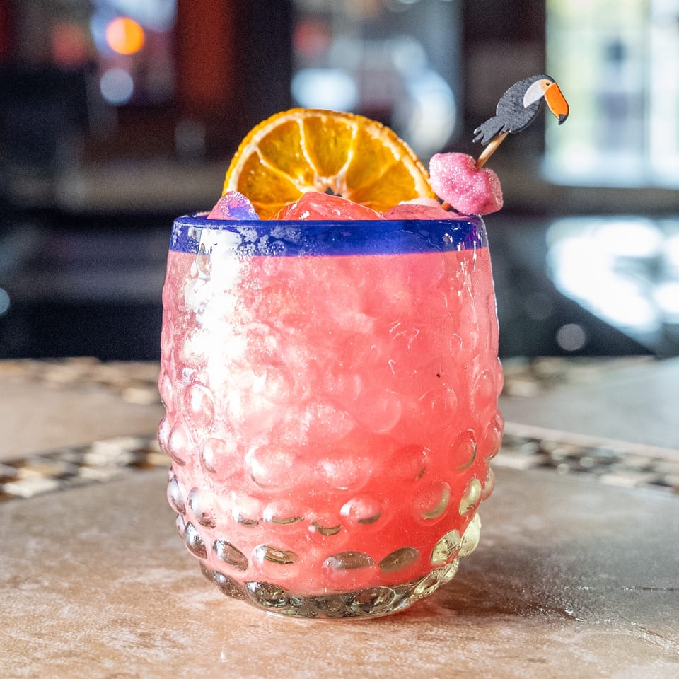 Pink frozen cocktail in a textured glass, garnished with an orange slice, berries, and a toucan pick.