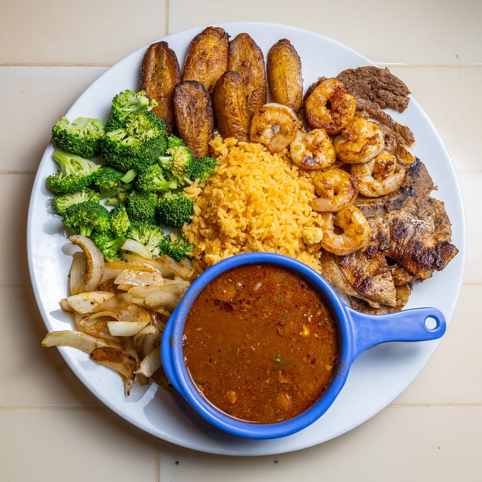 Plate of yellow rice, grilled shrimp and steak, fried plantains, broccoli, sautéed onions, and brown dipping sauce.