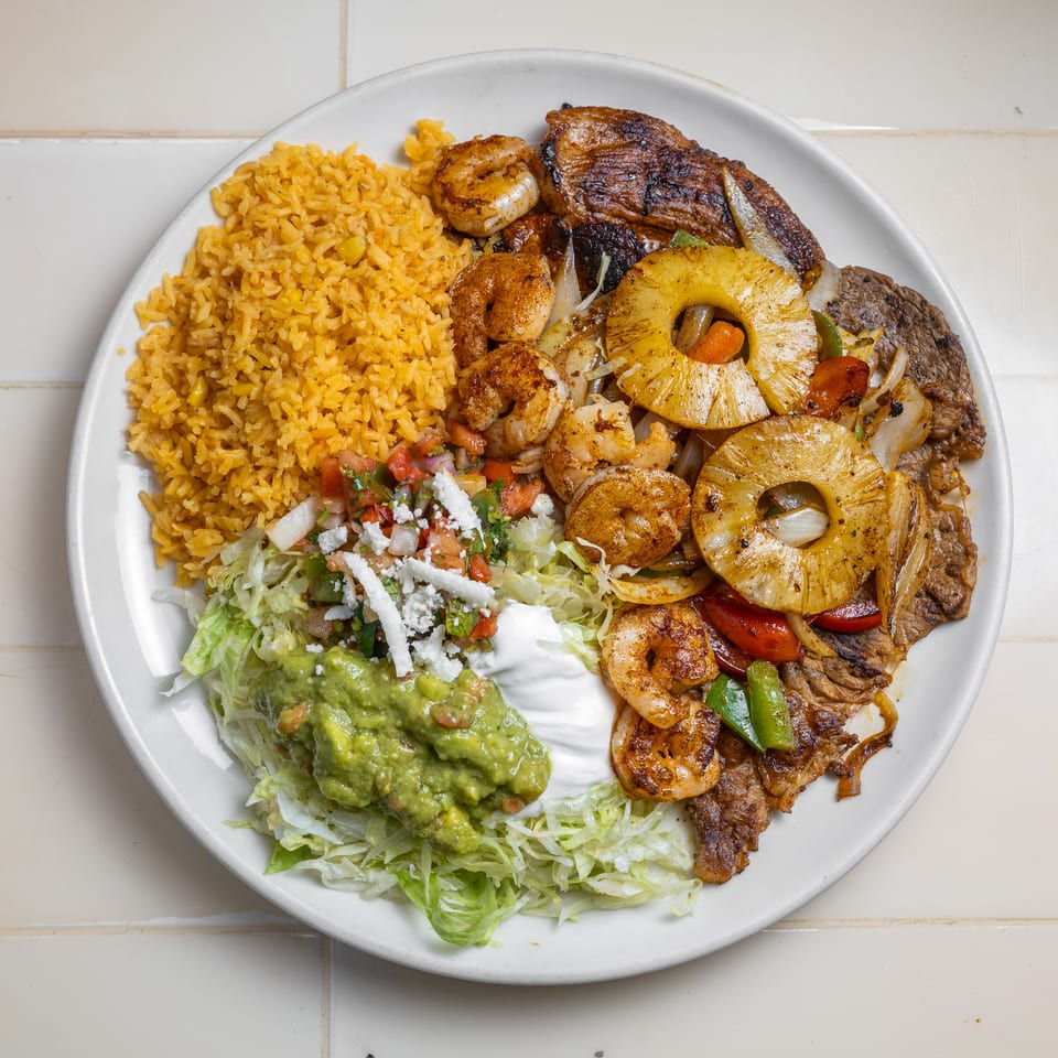 Plate of Mexican-style rice, grilled steak, shrimp, pineapple, vegetables, salad, guacamole, sour cream, and cheese.