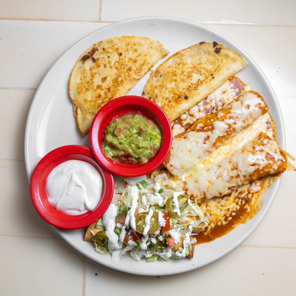Plate of cheesy enchiladas, toasted quesadillas, salad, guacamole, and sour cream on a tiled table.