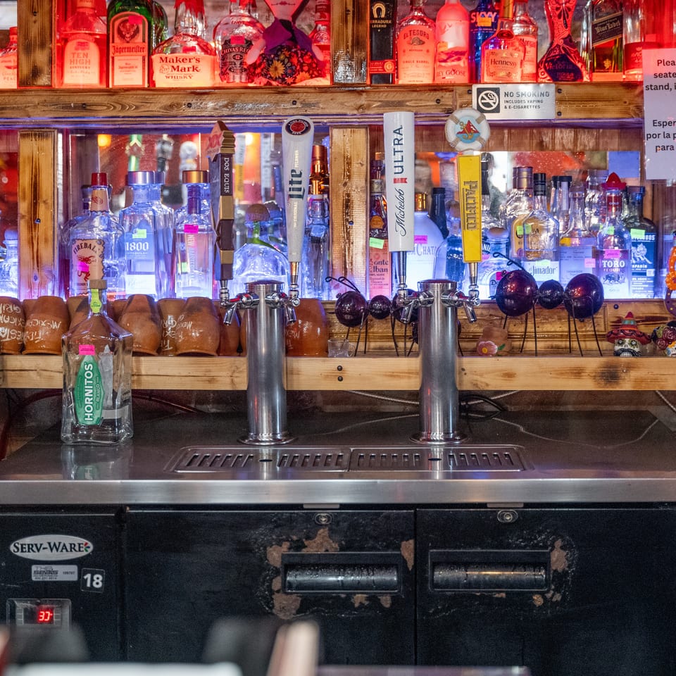 Colorfully lit bar displays liquor bottles, beer taps, mugs, and decorative skulls beneath a bilingual seating notice.