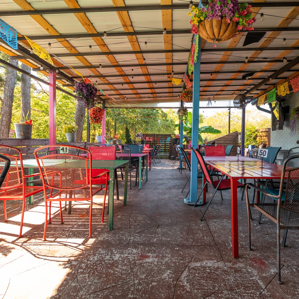 Colorful outdoor restaurant patio with metal tables, hanging flower baskets, decorative banners, and surrounding trees.