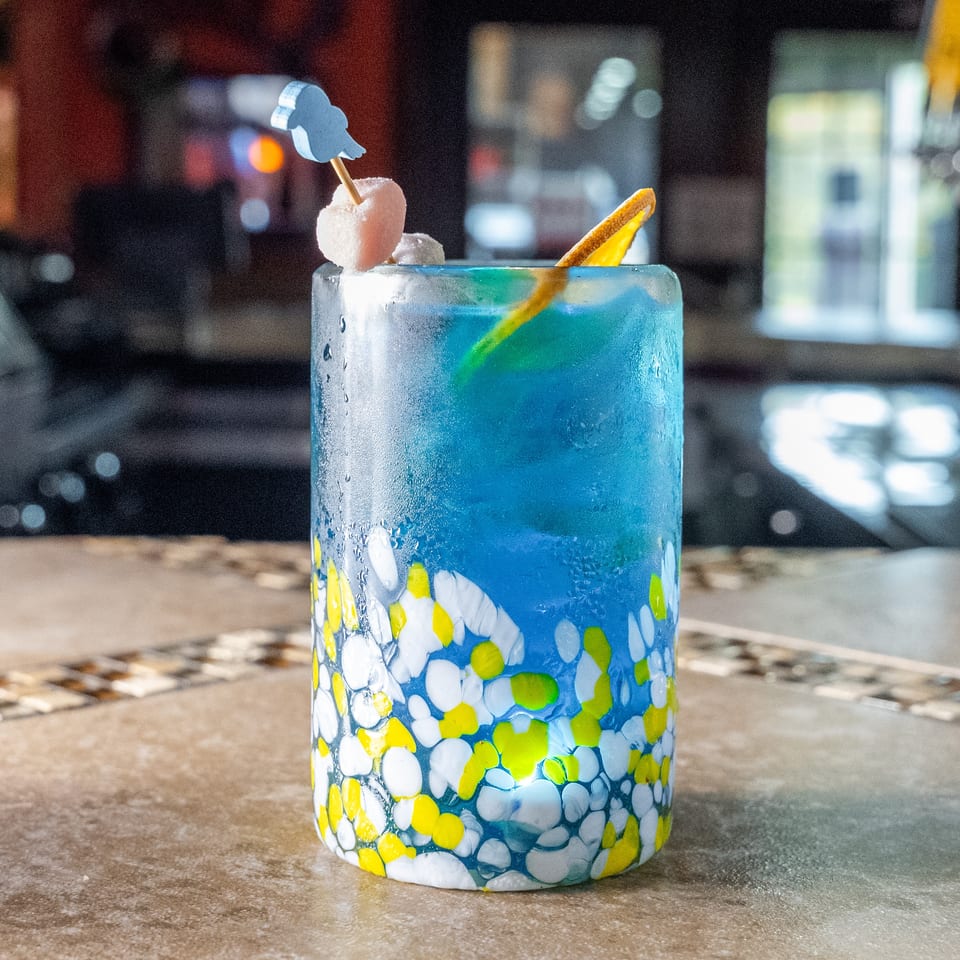 Bright blue cocktail in a colorful mosaic glass, garnished with citrus and a pink fruit skewer, sits on a restaurant bar.