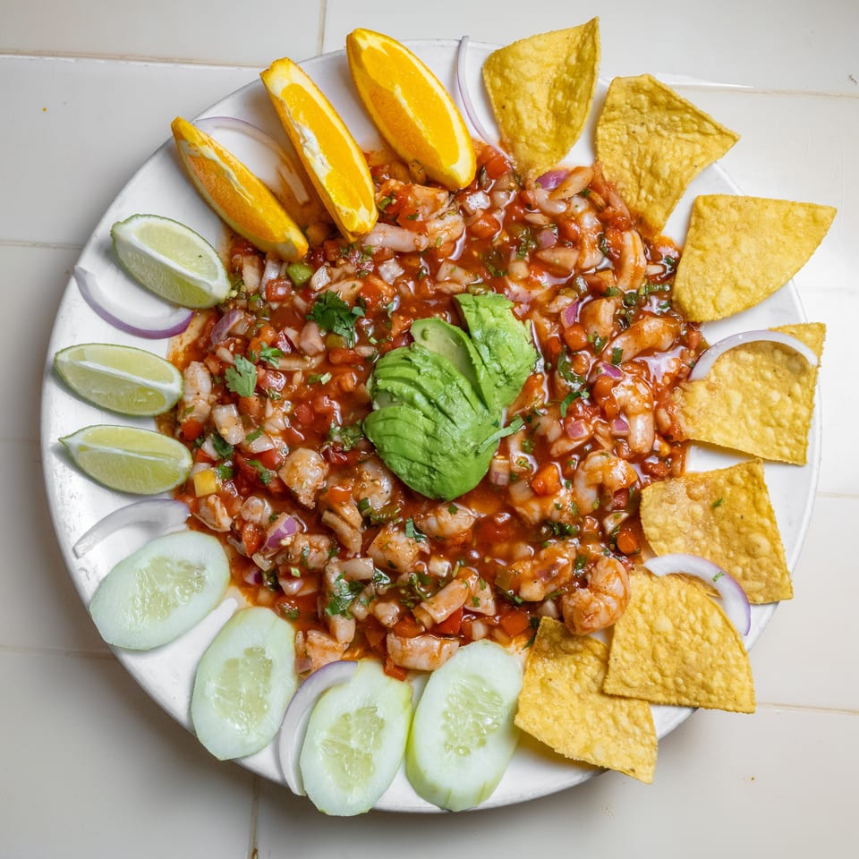 Shrimp ceviche with avocado, diced vegetables, lime, cucumber, orange wedges, red onion, and tortilla chips on a white plate.